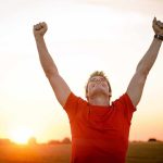 Man in a red shirt celebrating with arms raised against a sunset