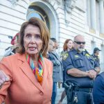 Trump UNLEASHES “Evil” Bomb on Pelosi Exit A woman in an orange blazer speaking passionately at a rally with security personnel in the background