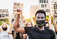 BLM Leader INDICTED – Facing 25 Counts! Protester raises fist, wears mask, signs in background.