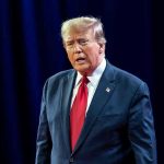 Man in suit with red tie speaking on stage.