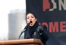 AOC Faceplants In Germany – Kamala 2.0 Woman speaking passionately at podium during outdoor event.