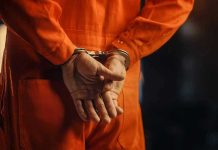 Authorities Discover Massive Fraud In Naturalization Tests – Ringleader Arrested Person in orange jumpsuit with handcuffs behind back.
