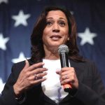Kamala Makes DESPERATE Move In Pathetic 2028 Push Woman speaking with microphone, American flag background.