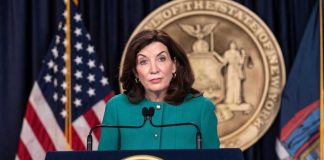 Governor CAUGHT! Paying Cops to Shield Illegals! Kathy Hochul speaks at podium with New York State emblem.