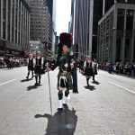 Participants in traditional Scottish attire marching in a parade on a city street