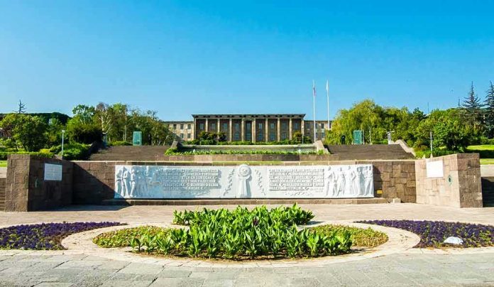 Government building behind landscaped gardens and a stone relief wall