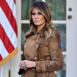Melania Trump standing in a brown suede coat near an American flag