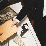 Two people exchange a manila envelope over a table with cash and a handgun