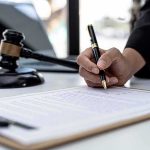 Migrant Maniac Kills 3 in Psychotic Attack Hand signing legal document beside gavel and laptop