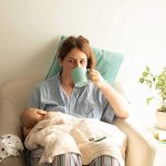 Mother nursing baby while drinking from a mug in a cozy chair