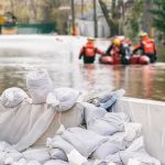 State of Emergency – Flash Flood Wrecks Region Sandbags stacked near a flooded street with rescue workers in a boat